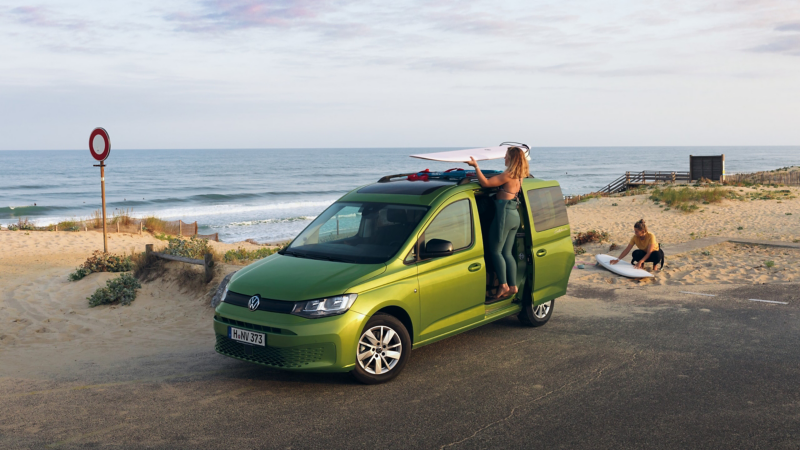 Ein Caddy California steht mit zwei Surferinnen am Strand.