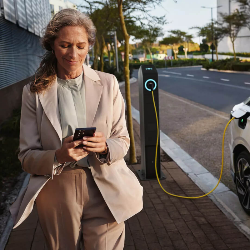 A smiling businesswoman in a beige suit looks at her smartphone while walking on a sidewalk, next to a public charging post connected to a white electric car via a yellow cable.