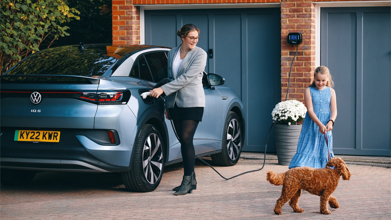 a woman plugging a light blue ID.5 into a home wallbox whilst a little girl plays with a scruffy brown dog