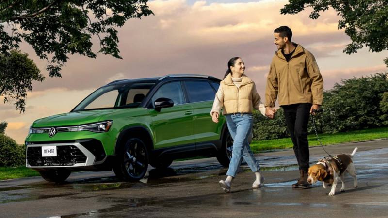Pareja camina, mientras el hombre sujeta una correa para perro. Detrás, un Volkswagen Taos de color verde.