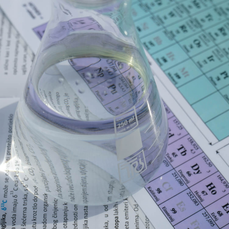 A glass beaker filled with clear liquid placed over a periodic table of elements, highlighting the chemical research behind LFP and sodium-ion batteries.