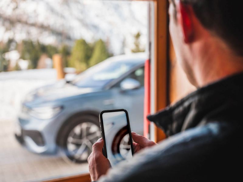 Un homme consulte les chaines à neige sur son smartphone, une Volkswagen ID. est visible par la fenêtre.