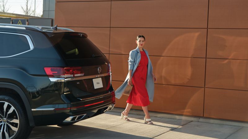 Une femme portant une robe rouge et un trench bleu pâle marche à côté d'un Atlas 2022 de Volkswagen noir profond nacré.