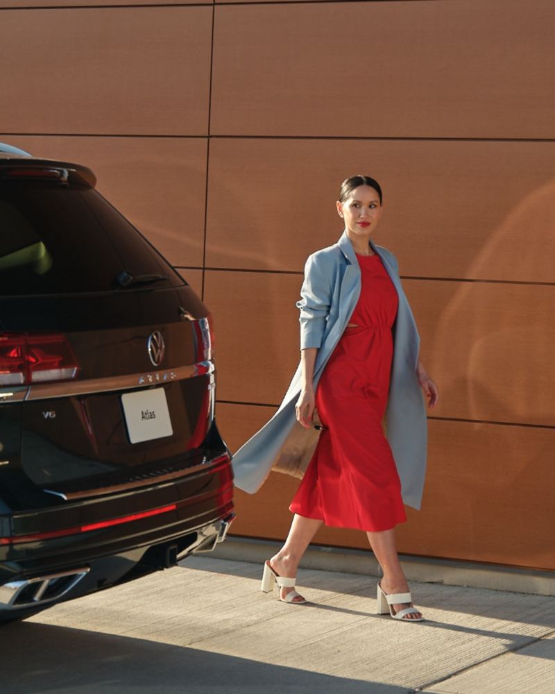A woman in a red dress and a light blue trench coat walking next to a Deep Black Pearl 2022 Volkswagen Atlas.