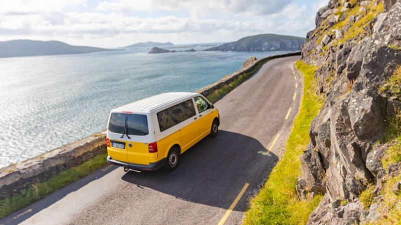 Van T6 jaune et blanc Volkswagen en circulation sur une route au bord de mer et en montagne.