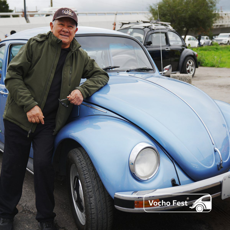 Conductor de Vocho posa sobre un Volkswagen sedán de color azul. 