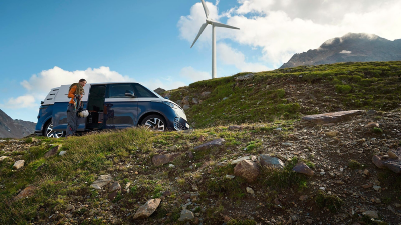 Una persona junto a una furgo Volkswagen en la montaña y un aerogenerador de fondo