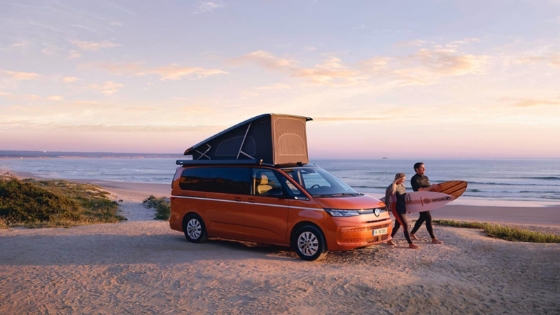 Dos personas con tablas de surf en la playa junto a una Volkswagen California