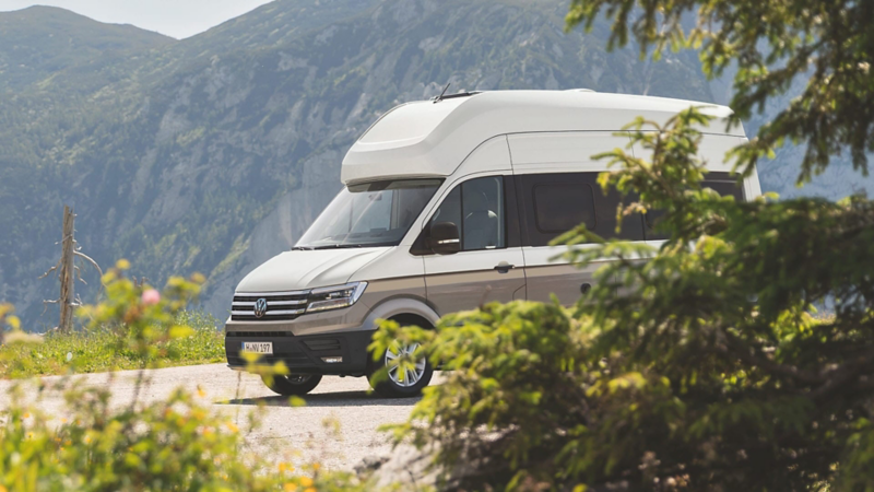 Vista lateral de un VW Grand California en una carretera boscosa. Detrás hay un paisaje montañoso.
