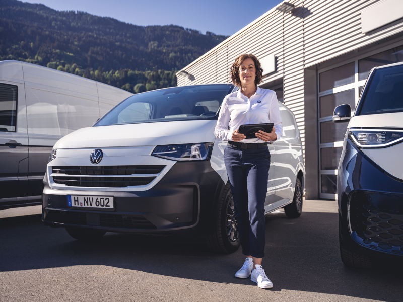 Una mujer vestida de Volkswagen con una tablet en la mano y unas furgonetas blancas detrás