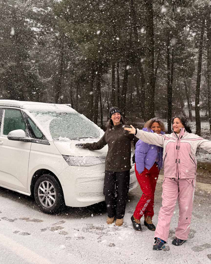 Tres personas posando felices con su furgo Volkswagen mientras nieva en un paisaje natural