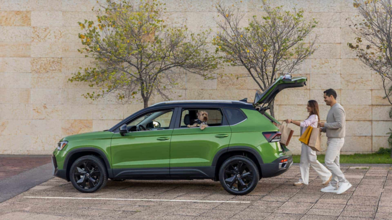 Pareja guarda equipaje en la cajuela de una SUV Taos de color verde, mientras el hombre carga a su perro.