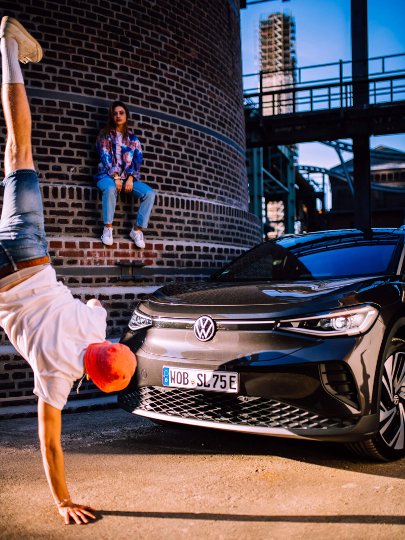 Un homme fait le poirier sur une main devant un Volkswagen ID.4, en arrière-plan, une jeune fille est assise sur les marches d'une tour de brique.