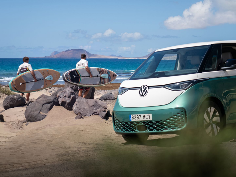 Unos surferos de espalda sujetando unas tablas junto una Volkswagen ID.Buzz verde y blanca parada junto a la playa