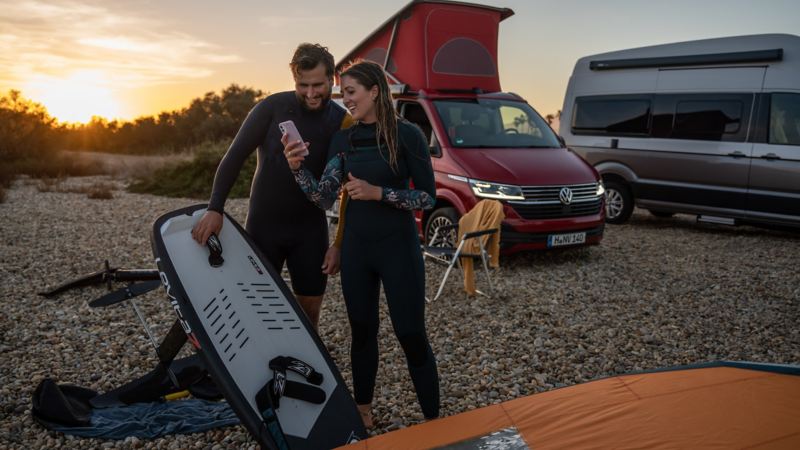 Two surfers are standing next to a VW California while looking at a mobile phone. 