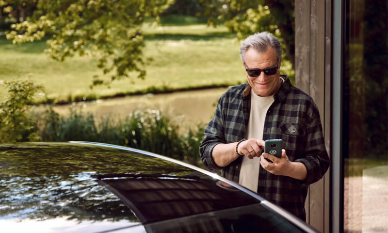 A smiling man wearing sunglasses and a plaid shirt looking at his smartphone next to a car roof, with a natural landscape in the background.