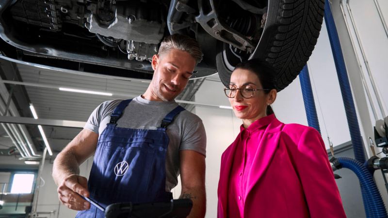 Un technicien Volkswagen et une femme discutent sous un véhicule présent dans l'atelier.