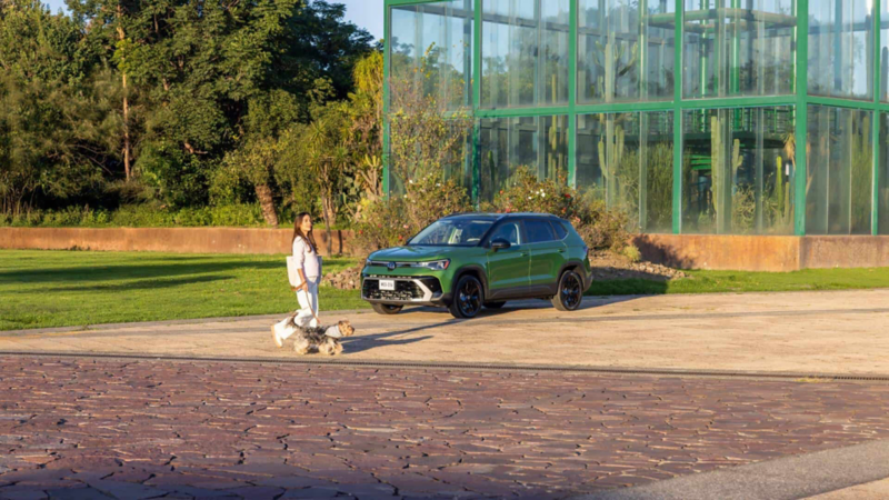 Mujer pasea a su perro frente a Volkswagen Taos de color verde estacionado junto a edificio de cristal.