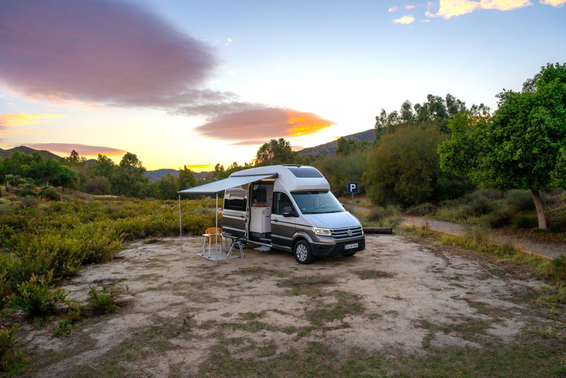 Un Grand California Volkswagen est garé dans un paysage naturel, sous un ciel de coucher de soleil. Son auvent est déplié et une table et des chaises de camping sont installées à l'extérieur, à côté de la porte ouverte du véhicule.