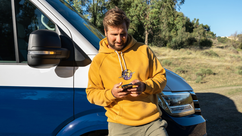 Mann lehnt entspannt am VW California und blickt auf sein Smartphone.