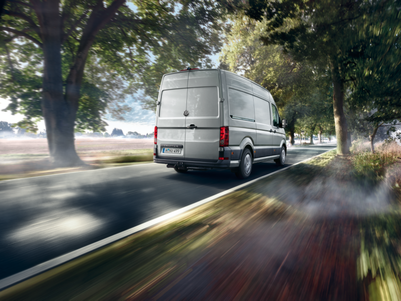 Volkswagen Crafter Furgon jedzie po drodze asfaltowej między drzewami.