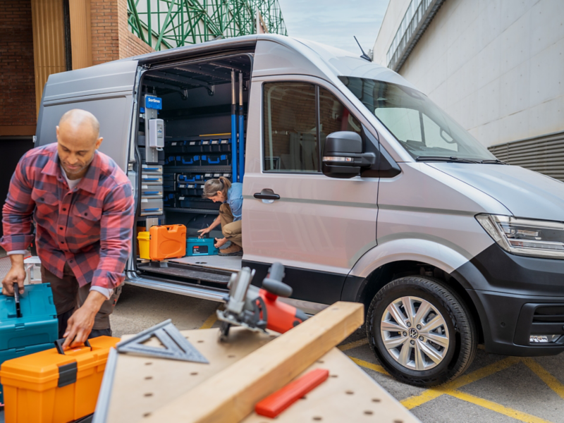 Ein zur mobilen Werkstatt ausgebautem VW Crafter von der Seite, mit offenen Seitentür, ein Handwerker arbeitet daneben an einem Tisch.