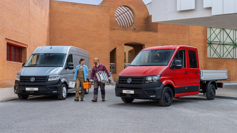 Two workers stand between a silver Volkswagen Crafter van and a red Crafter truck, chatting cheerfully. They carry tools, suggesting a workday setting.