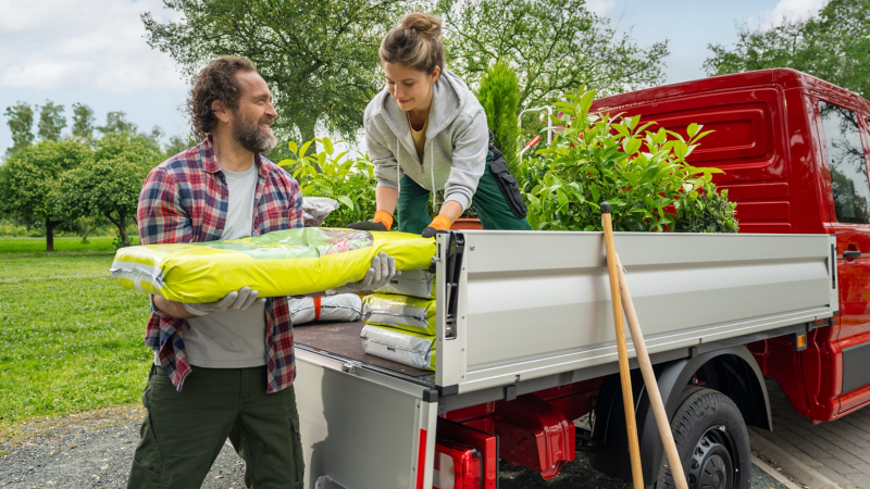 Zwei Gärtner beladen einen roten VW Crafter Pritschenwagen mit Erde und Pflanzen in der Natur.