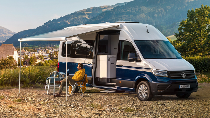 Der Volkswagen Multivan auf einer Wiese vor einer Stadt an den Bergen mit Camping-Funktion, eine Person sitzt auf einem Campingstuhl