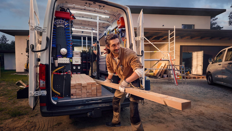 Ein Bauarbeiter lädt eine lange Holzplanke in das Innere eines weißen Transporters, dessen Hecktüren weit geöffnet sind und spezielle Regalsysteme im Laderaum sichtbar machen.