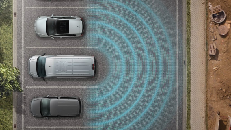 Foto toont een Volkswagen Bedrijfsvoertuigen Crafter op een parkeerplaats tussen twee andere auto's vanuit vogelperspectief. Blauwe halve cirkels geven aan hoe de optionele Blind Spot Sensor incl. uitparkeerhulp werkt.