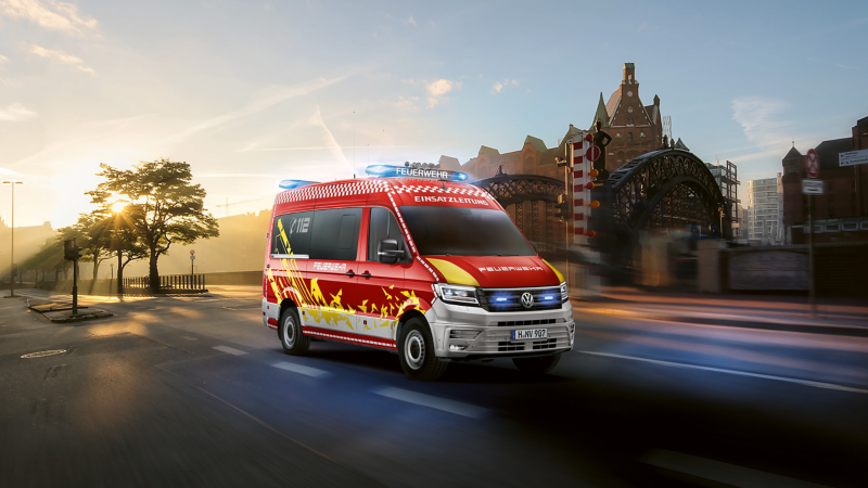 Un véhicule de commandement des pompiers, un Volkswagen Crafter rouge, est en circulation sur une route en ville, avec le soleil levant en arrière-plan.