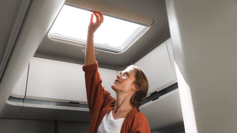 A woman is opening the skylights in the Grand California in the morning and lets sunlight into the mobile home.