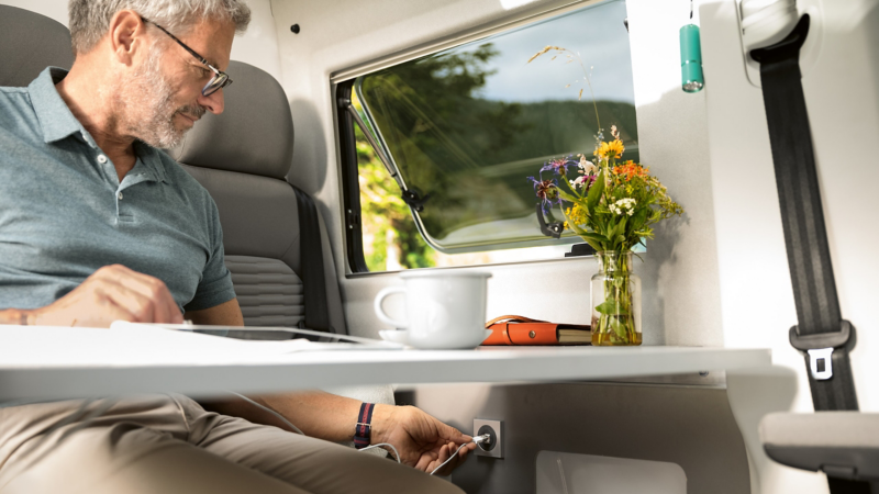 A man is sitting a table and is currently plugging his tablet’s charging cable in the socket in the vehicle wall. 