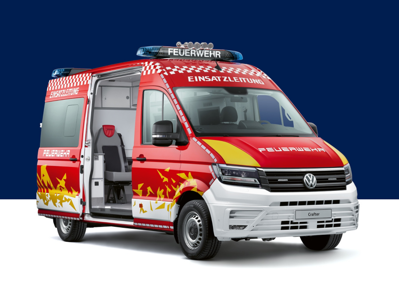 Un Volkswagen Crafter aménagé en véhicule de commandement pour les pompiers (inscription "Feuerwehr Einsatzleitung" en allemand). Le véhicule est rouge et blanc, avec des gyrophares bleus sur le toit. Sa porte latérale coulissante est ouverte, laissant entrevoir l'intérieur.
