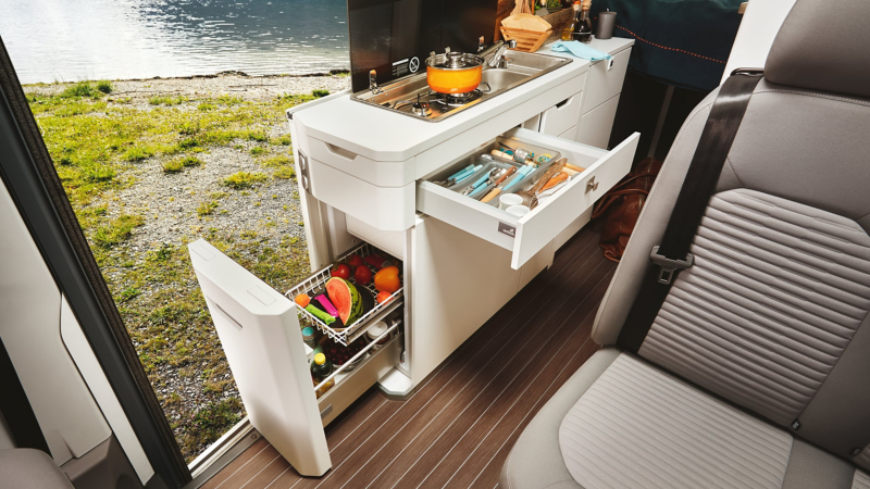 We see the kitchen area in the Grand California. A pan is standing on the two-hob gas stove. The open sliding door reveals a view of a mountain lake.