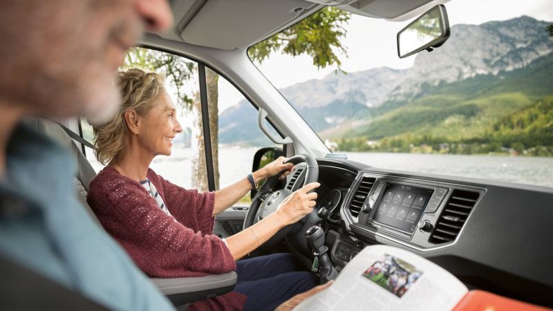 Widok od strony pasażera obok kierowcy do kokpitu. Kobieta siedzi za kierownicą, jej mąż na miejscu pasażera. W tle widać panoramę górską z jeziorem.