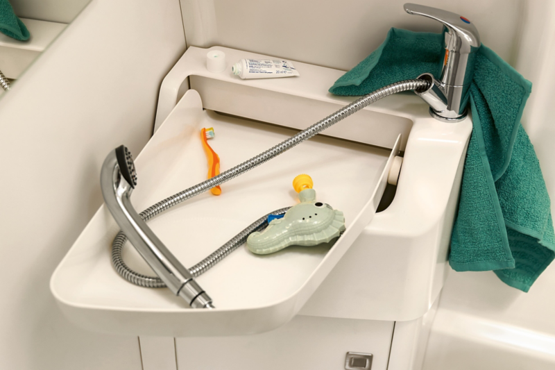 A close-up of the bathroom in the Volkswagen Commercial Vehicles Grand California. The flexible wash basin is folded out. It contains a shower head, a children’s toothbrush and a colourful toy.