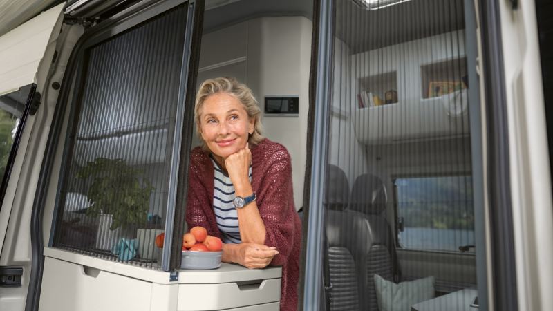 Una donna, appoggiata all’armadietto della cucina, guarda attraverso la porta scorrevole aperta. Sui lati destro e sinistro sono presenti le zanzariere. 
