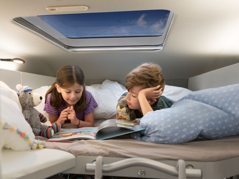 Två barn myser i loftsängen under takfönstret i en VW Grand California husbil.