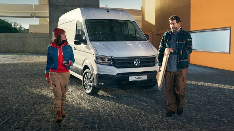Bildet viser en hvit stor Volkswagen Crafter varebil som står parkert foran en orange vegg og en dame som står foran kassebilen