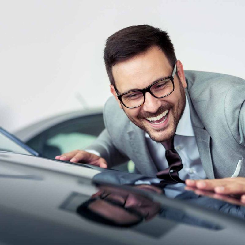 Hombre con lentes toca el cofre de un carro que compró en concesionaria. 