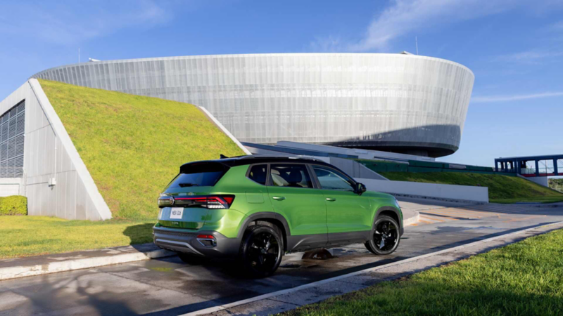 Taos VW verde por la parte trasera junto a un edificio moderno de formas curvas y techo con césped.