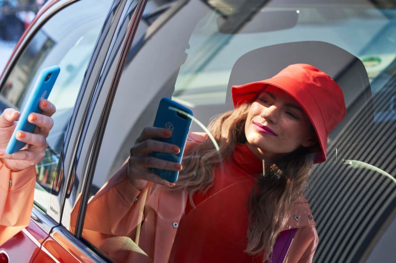 Une femme utilise son smartphone devant la portière de sa voiture Volkswagen et nous voyons son reflet dans la vitre.
