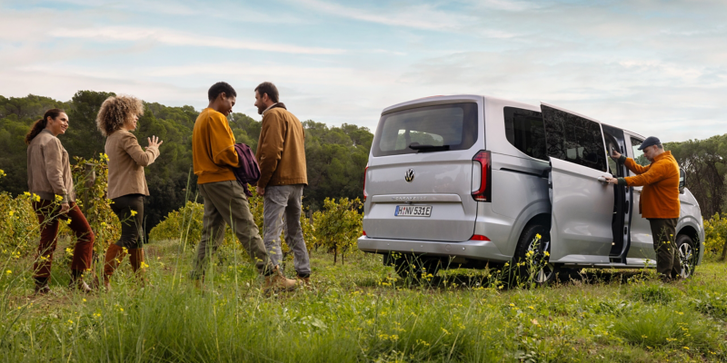 Der VW Caravelle parkt auf einer Wiese und vier Personen stehen dahinter.