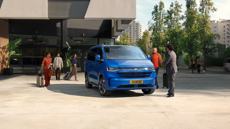 Un van Volkswagen bleu vif est garé devant un bâtiment moderne avec des personnes autour, certaines avec des bagages. Au premier plan à droite, deux hommes conversent près du véhicule.