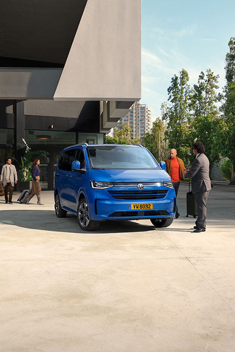 Image montrant le nouveau monospace Volkswagen Multivan T7 de couleur bleue garé devant un bâtiment moderne. Trois hommes et une femme se tiennent autour du véhicule avec des bagages. Le bâtiment est entouré d'arbres.