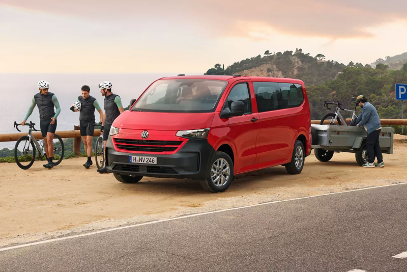 Une VW Caravelle rouge est garée sur un accotement avec une remorque à laquelle est attaché un vélo et trois personnes passent devant la Caravelle.