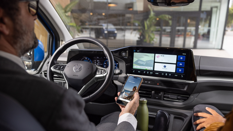 Eine Person am Steuer eines parkenden VW Transporter blickt auf ein Smartphone mit geöffneter VW Connect App.