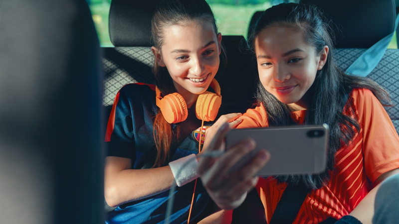 Zwei Mädchen sitzen auf der Rückbank und gucken sich etwas über ein angestecktes Smartphone an.